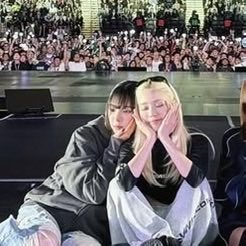 Two young women pose together at a concert, with one making a heart shape with her hands near the other's face, while a large crowd fills the background.
