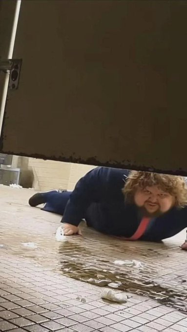 A person with curly hair and a beard crawling on the ground beneath a heavy door, with a spilled drink and cup visible on the tiled floor.