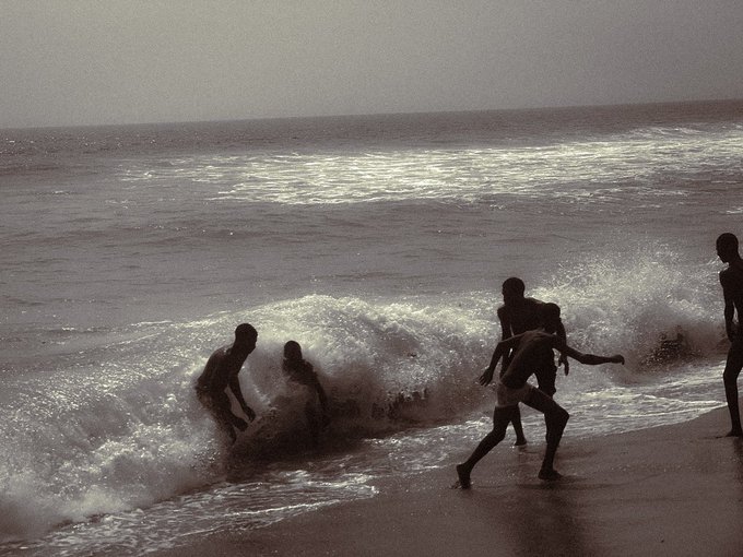 A vintage-style photograph showing the silhouettes of several young men playing in the crashing surf at a beach during what appears to be sunset or sunrise.
