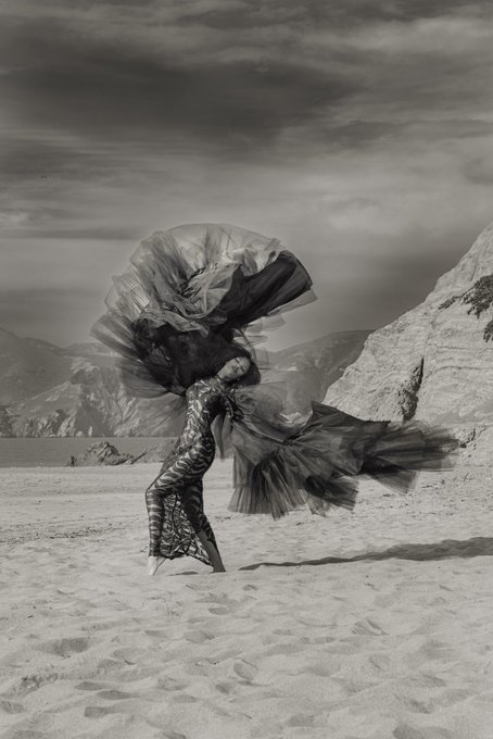 A black-and-white photograph shows a figure in a flowing, voluminous dress posing on a sandy shore against a mountainous landscape.