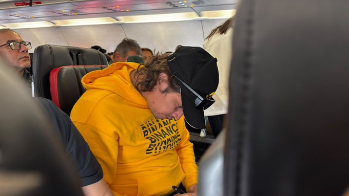 A passenger wearing a yellow "RISE AND GRIND" hoodie sleeps with his head tilted forward on an airplane.