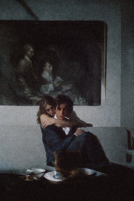 A moody photograph captures a young couple sitting closely together on a sofa beneath a large classical painting.