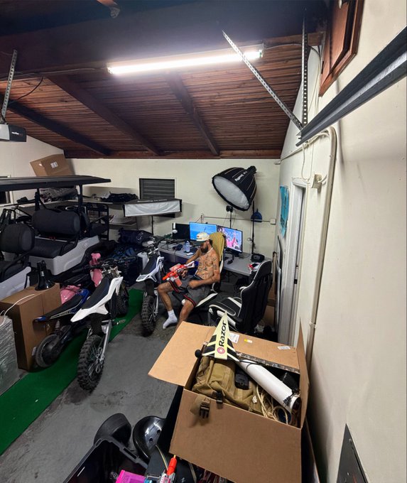 A cluttered garage interior with two dirt bikes, a golf cart, photography lighting equipment, and a person sitting at a computer desk in the back.