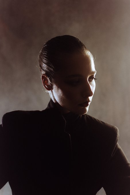 A moody, low-key portrait of a woman with slicked-back hair, wearing a black high-neck garment, illuminated by soft side lighting against a textured wall.