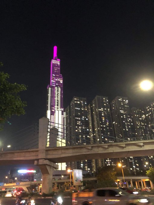 A nighttime photograph of a city skyline featuring a tall, illuminated skyscraper glowing with purple lights, with apartment buildings, an overpass, and the moon visible in the sky.