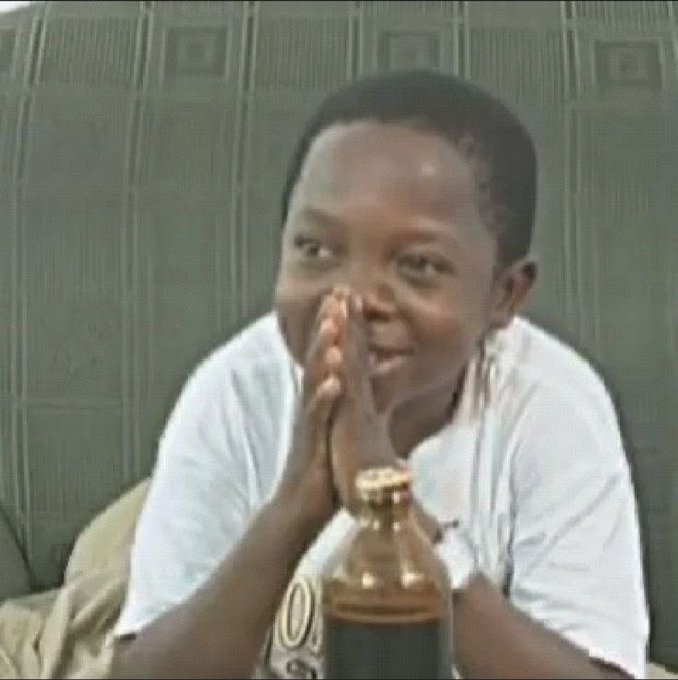 A young man in a white t-shirt sits on a couch with his hands clasped together in a prayer-like gesture, with a glass bottle in front of him.