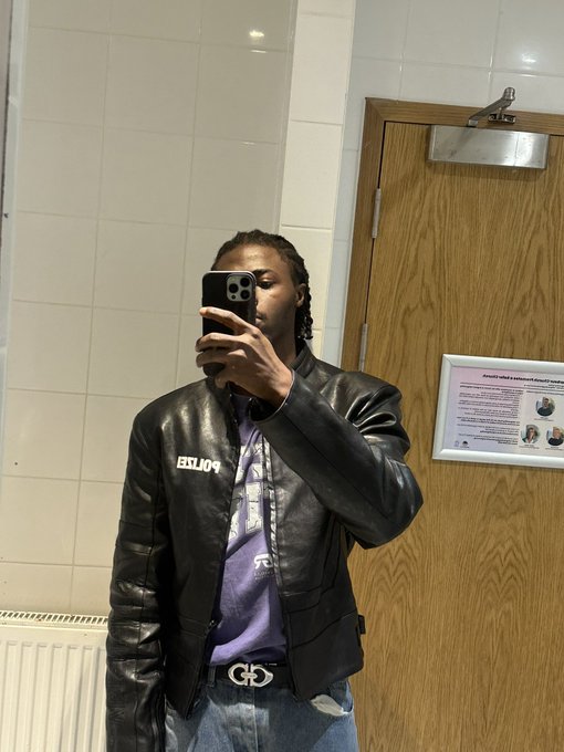 A mirror selfie of a person wearing a black leather POLAR jacket over a purple shirt and blue jeans with a designer belt, taken in what appears to be a public restroom.