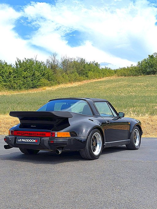 A black Porsche 911 (likely a 930 Turbo 'Widowmaker') parked on pavement with a grassy hill and trees in the background, featuring a 'LE PADDOCK' license plate.