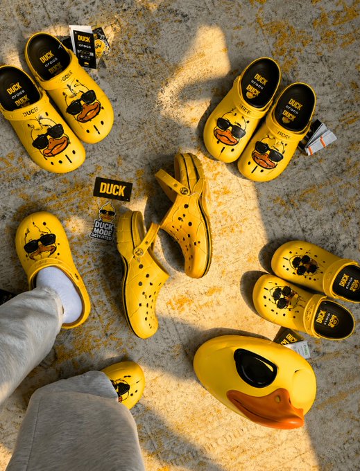 Multiple pairs of yellow Crocs with 'Duck Mods' accessories, featuring sunglasses and beak attachments, are arranged on a speckled floor.