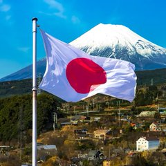 A Japanese flag waves in the foreground with snow-capped Mount Fuji and a town visible in the background under a blue sky.