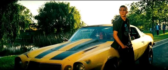 A cinematic widescreen still showing a young man leaning against a classic yellow muscle car with black racing stripes, parked near a pond in a green, sunlit park setting.