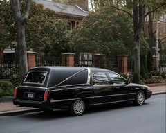 A black hearse is parked along a residential street lined with trees and houses, visually echoing the post's joke about the stock $HTZ "coming out of the hearse" after short sellers are finished with it. The macabre humor plays on the vehicle's funeral connotations as a metaphor for the stock's struggles.