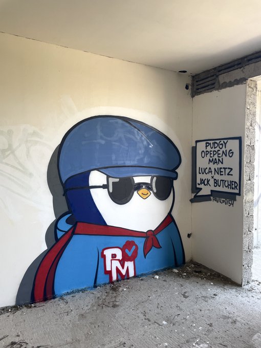 A street art mural of a blue penguin character wearing sunglasses, a blue beanie, and a red cape with 'RM' lettering, alongside a speech bubble reading 'PUDGY OPEPENG MAN LUCA NETZ JOCK BUTCHER' signed by 'PALVUTOPIA'.