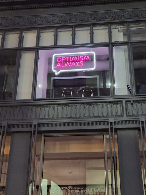 A neon sign in a speech bubble reading 'OPTIMISM ALWAYS' in pink, displayed in the window of a building with classical stone facade architecture.