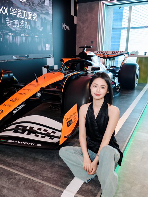 A woman crouching next to an orange and black Formula 1 race car on display, with a screen behind showing Chinese text for a 'KIX 华语见面会' event in Singapore.