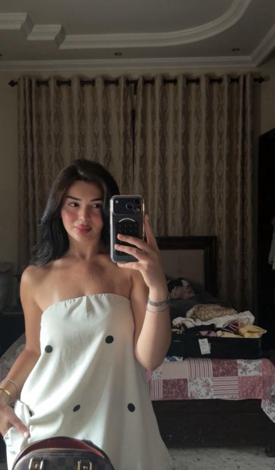 A mirror selfie of a young woman wearing a white strapless dress with black polka dots, standing in a bedroom with brown curtains.