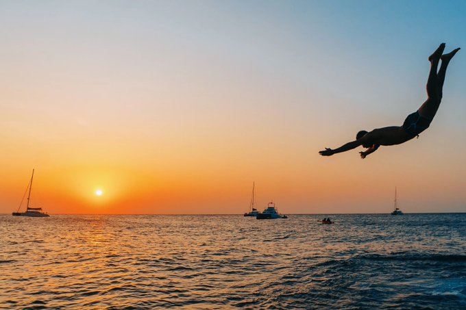A silhouetted person dives through the air over the ocean at sunset, with sailboats visible on the horizon and the sun setting in a warm orange sky.