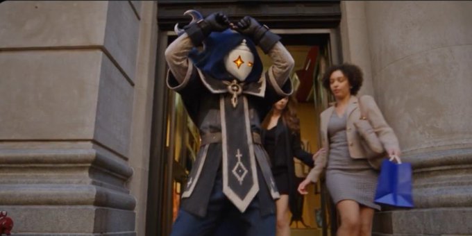 A character in blue and silver assassin-style robes pulling up a hood walks out of a building past people on the street.