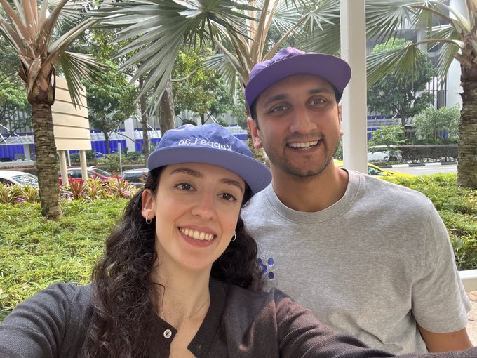 A smiling couple takes a selfie outdoors with palm trees in the background; the woman wears a blue cap with the text "Alexis Sablone".