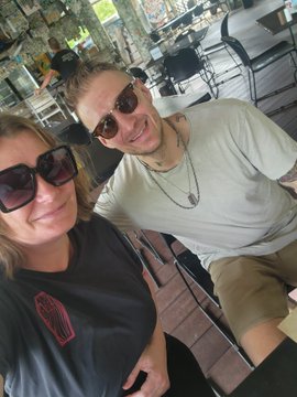A selfie of a woman with sunglasses and a man with sunglasses smiling together, appearing to be at an outdoor venue.