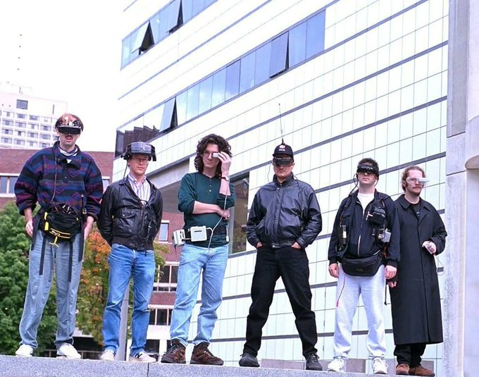 A diverse group of seven individuals stands outside a modern glass building, wearing an array of tech accessories including VR/AR headsets, a large belt-mounted computer, and various wearable devices—evoking a retro-futuristic or early-90s vision of wearable computing that contrasts humorously with the post's commentary about AI context lookups and modern model differentiation.