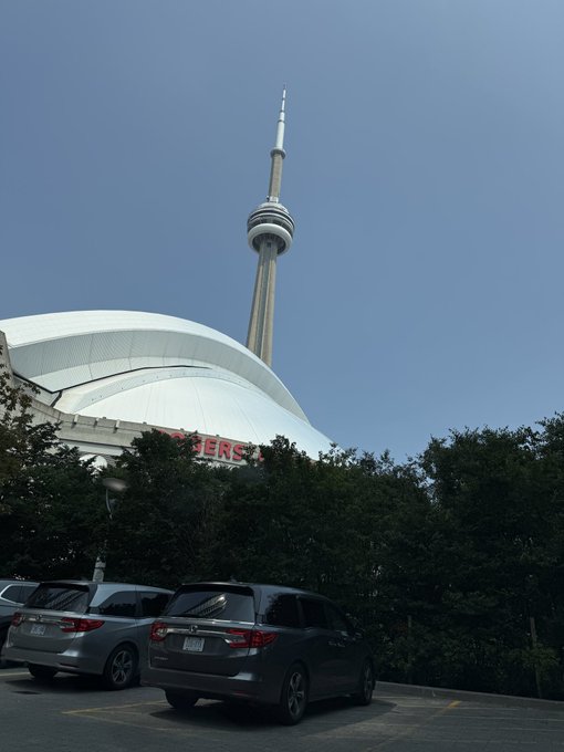 The Rogers Centre (with 'ROGERS' visible on the facade) in Toronto with the CN Tower rising into a clear blue sky, with parked cars and trees in the foreground.