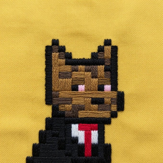 An embroidered pixel art design on yellow fabric depicts a brown and black cat character wearing a white shirt with a red tie, resembling a Minecraft skin.