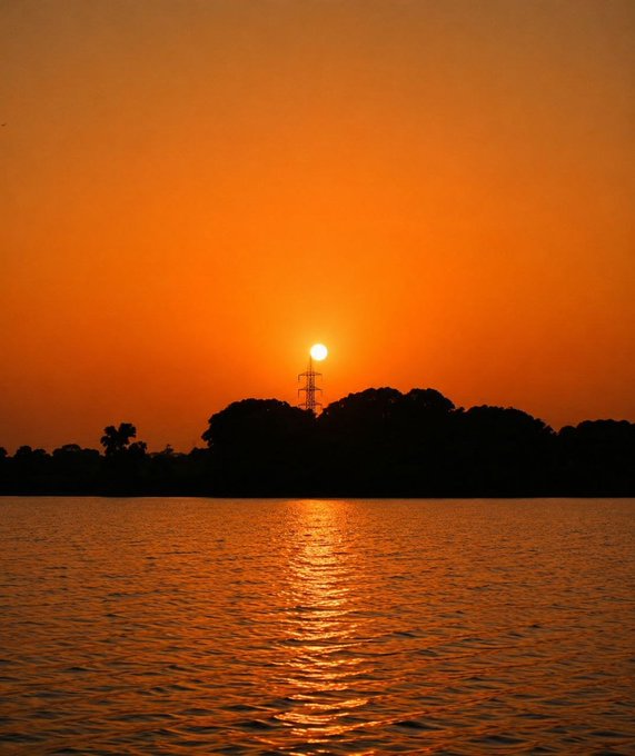 A serene sunset over a body of water, with the sun glowing orange behind silhouetted trees and a transmission tower.