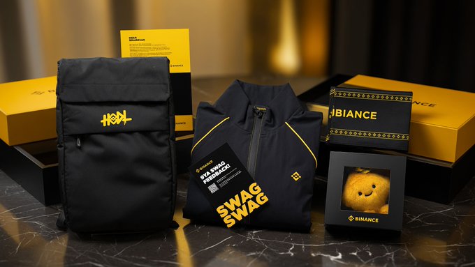A collection of Binance-branded merchandise including a black backpack, a black and yellow jacket, packaging boxes, and a plush Binance mascot toy displayed on a dark surface.