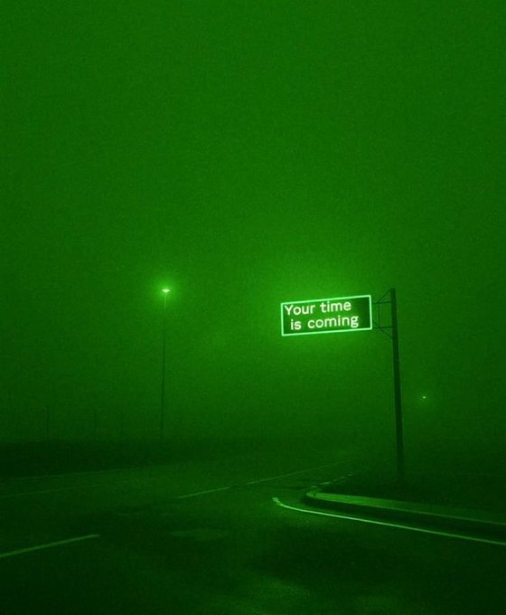 A green-tinted night photograph of a foggy street with a streetlight and a road sign that reads 'Your time is coming'.