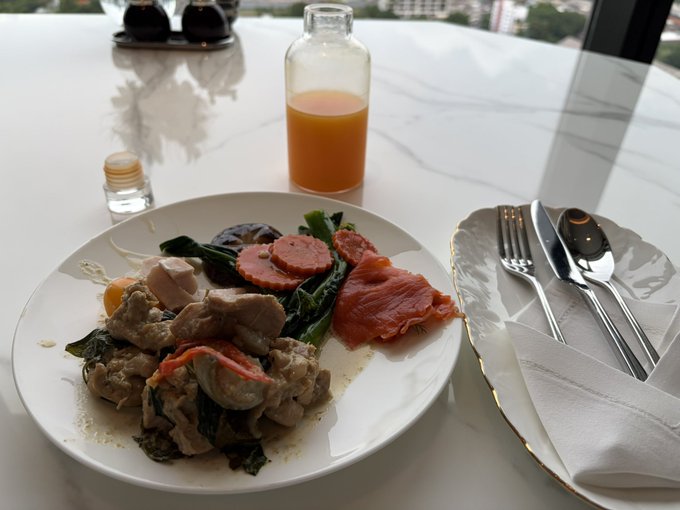The image shows a white plate with food, including meat, vegetables, and an orange drink placed on a dining table. There are also silverware items such as forks and spoons nearby. The background of the photo features a window overlooking a cityscape, giving the scene a sense of openness and spaciousness. The post caption reads "午餐30u nice！", which is likely an advertisement for a restaurant or food establishment in the area.