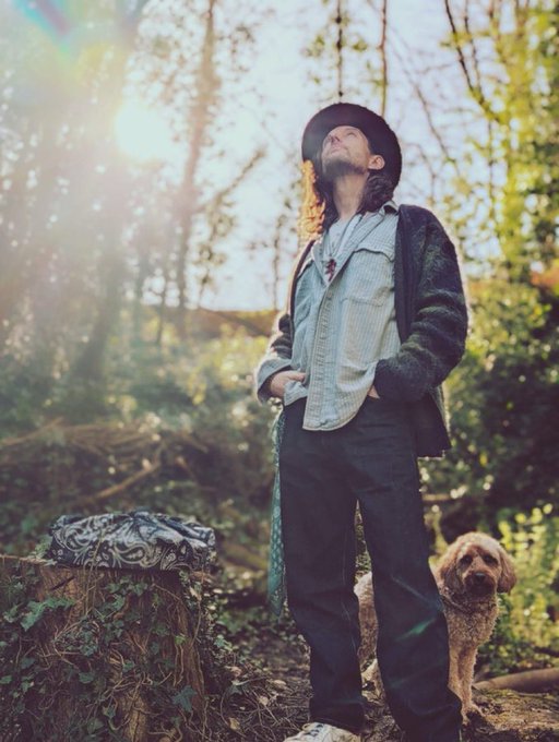A man in a bucket hat, jacket, and dark pants standing in a forest looking up at the sun, with a curly-haired dog beside him.
