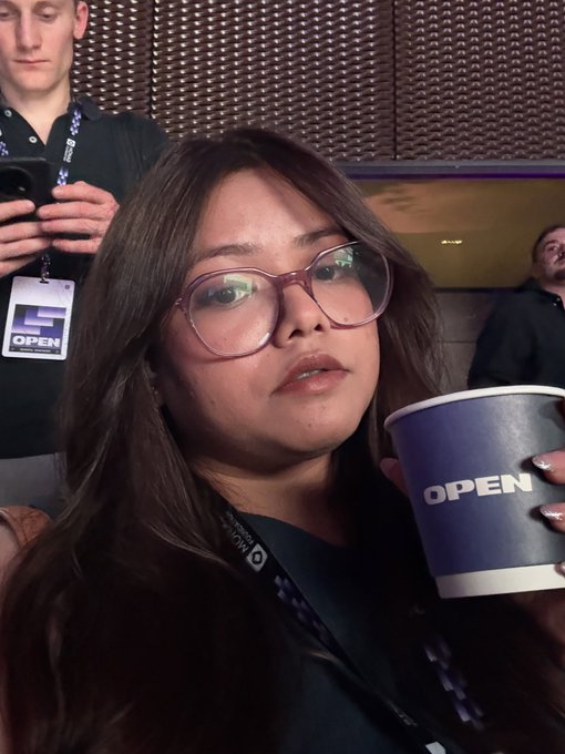 Selfie of a woman wearing glasses holding a blue 'OPEN' branded cup at what appears to be an 'OPEN AI' event, with another attendee visible behind her.