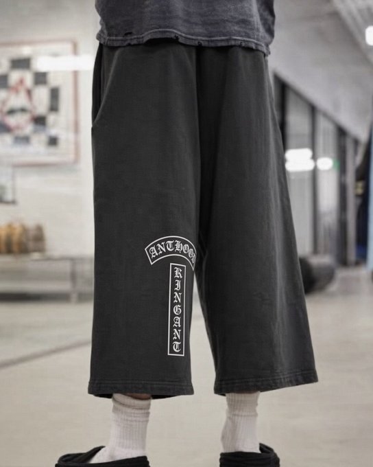 A person models black wide-leg cropped pants featuring a white cross graphic with "ANTHOOD" arched at the top and "KINGDOM" written vertically down the cross. The streetwear piece visually reinforces the post's promotion of AntHood, showcasing the apparel's distinctive branding as part of their brand's identity and community message.
