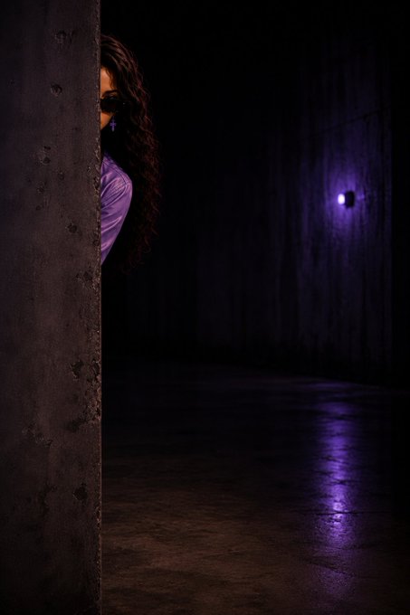 A moody, dark photograph of a woman with long hair peeking out from behind a concrete wall, illuminated by a purple light in an otherwise shadowy space.