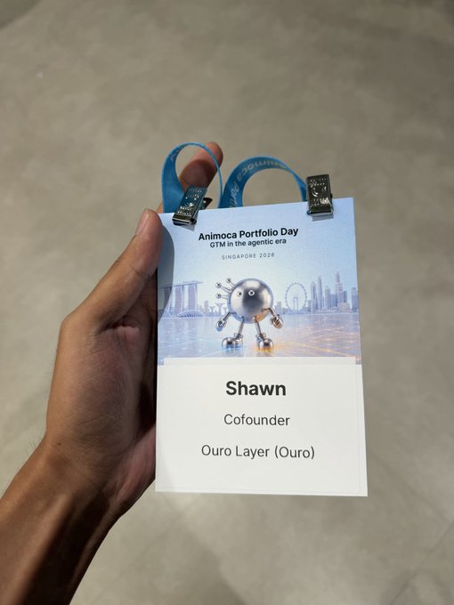 A hand holding an 'Animoca Portfolio Day' attendee badge for 'Shawn, Cofounder, Ouro Layer (Ouro)' from Singapore 2026, featuring a metallic robot character on the lanyard.