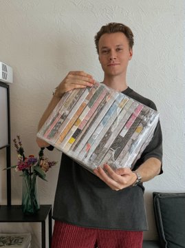 A young man wearing a dark t-shirt and red striped pants holds up a stack of books wrapped in plastic.