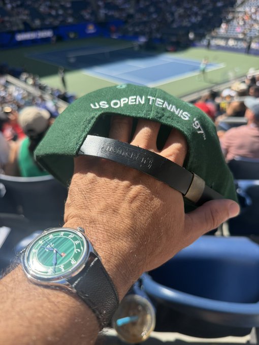 A person's hand, wearing a green-faced watch with 'H. Moser & Cie.' text and a beaded bracelet, holds up a green 'US OPEN TENNIS 197' cap while overlooking a tennis court.