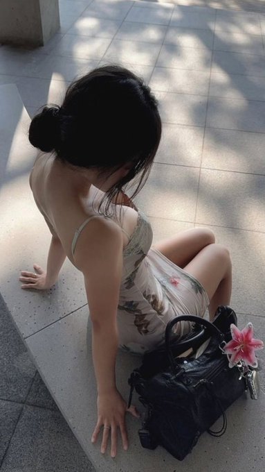 A woman in a floral dress sits on tiled pavement beside a black handbag adorned with a pink lily, while dappled sunlight filters across the scene. The contemplative, casual pose reflects the post's theme of pausing to consider diversification in the booming AI crypto sector rather than rushing in blindly.