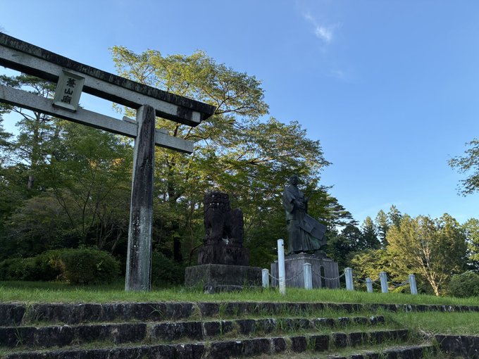A traditional Japanese stone torii gate (with the characters '愛宕') standing on grass next to stone statues of a komainu lion-dog and a seated figure, with green trees and a blue sky in the background.
