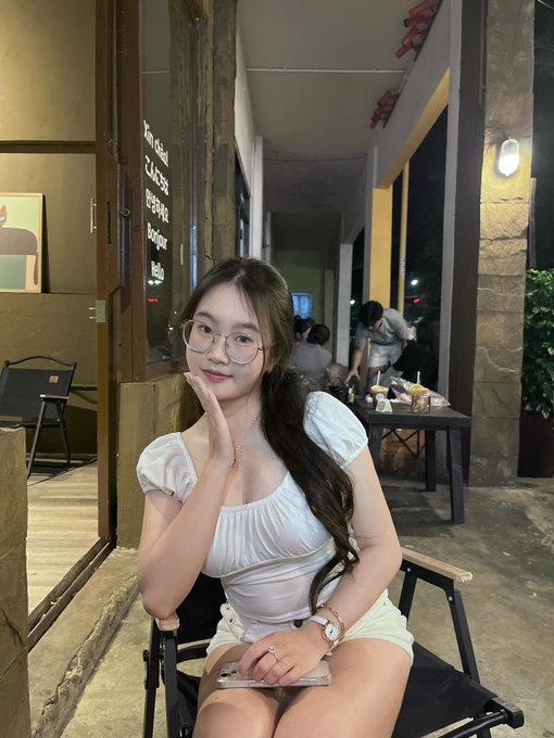 A young woman with long dark hair and glasses sits on a chair in a white puff-sleeve top and shorts, posing with her hand on her cheek at what appears to be an outdoor café at night.