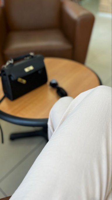A first-person view of someone's legs in white pants resting near a small wooden table holding a black handbag and sunglasses, with a brown armchair in the background.