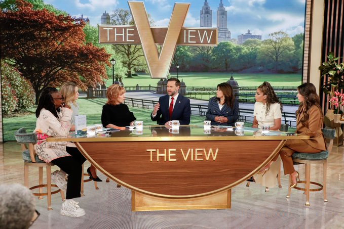 The image shows a group of six women sitting around a large wooden table, engaged in conversation with each other. They are all dressed up, suggesting that they might be attending an event or gathering. The table has a sign on top that reads "the view", indicating that this is likely a television show or news program being broadcasted at the time. One of the women appears to be wearing a tie, further emphasizing the formal nature of the occasion. A potted plant can also be seen in the background, adding some greenery and decoration to the scene. The overall atmosphere seems to be one of camaraderie and shared interest among the group members as they discuss various topics or events.