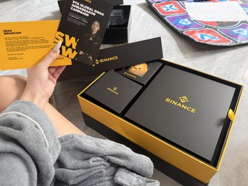The image shows a person displaying Binance 9th anniversary merchandise, featuring a black and gold "BINANCE" branded box containing a small black pouch and promotional cards with text like "THE BLOCKCHAIN CHANGING THE WORLD" alongside a round pin. The caption from the post reflects excitement about receiving the anniversary merch, specifically mentioning a jacket and backpack—though only the boxed items are visible in this view.