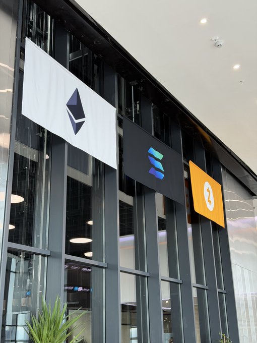 Three large hanging signs in a modern glass-walled building displaying the Ethereum logo on white, the Solana logo on black, and a third logo on an orange background.