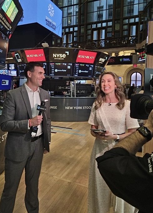 Two smiling news anchors in business attire stand inside the New York Stock Exchange trading floor, being filmed by a cameraman.