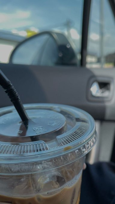 A close-up view of an iced coffee in a clear plastic cup with a black straw, held inside a car with the passenger window showing a bright outdoor scene.