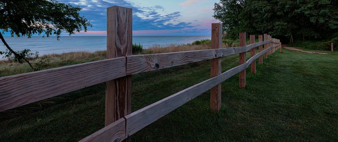 A wooden split-rail fence runs along a grassy bluff overlooking a large body of water under a dusky twilight sky.