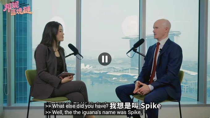 An interview shows Coinbase CEO Brian Armstrong seated with a female host in front of a window overlooking Singapore, with subtitles reading "What else did you have? I think it was called Spike" and "Well, the iguana's name was Spike, I" — referencing Armstrong's reveal of his pet iguana's name, which the post humorously promotes as the "$SPIKE" token linked to his known fondness for the name.