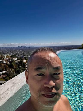 A selfie of a man in an infinity pool with a city skyline visible in the distance under a clear blue sky.